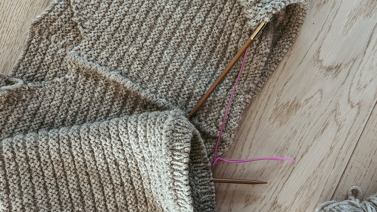 Bol og ermer i gråbeige ribbestrikk på pinnen, liggende på et lyst tregulv · ribbene ligger jevne
