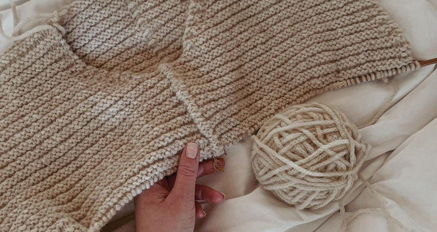 En hånd holder et påbegynt plagg i beige ribbestrikk ved siden av et nøste · ribbene har et rolig, jevnt mønster