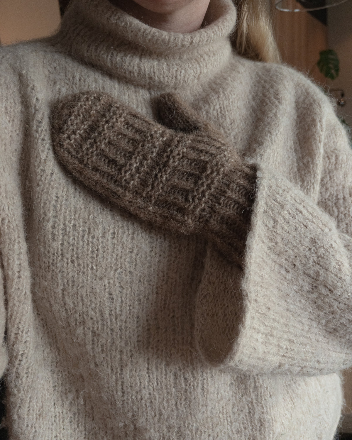 Mørkebrun Coffee Mitten holdt mod en cremehvid sweater · tommelfingerhullet markeres med en tråd undervejs