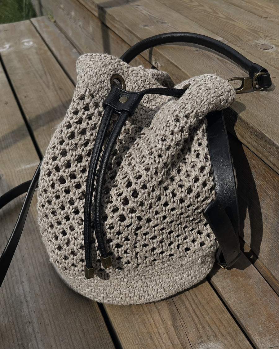 Bucket bag i netmønster i naturfarvet hør på trægulv, med sorte læderstropper · strikkes uden sømme