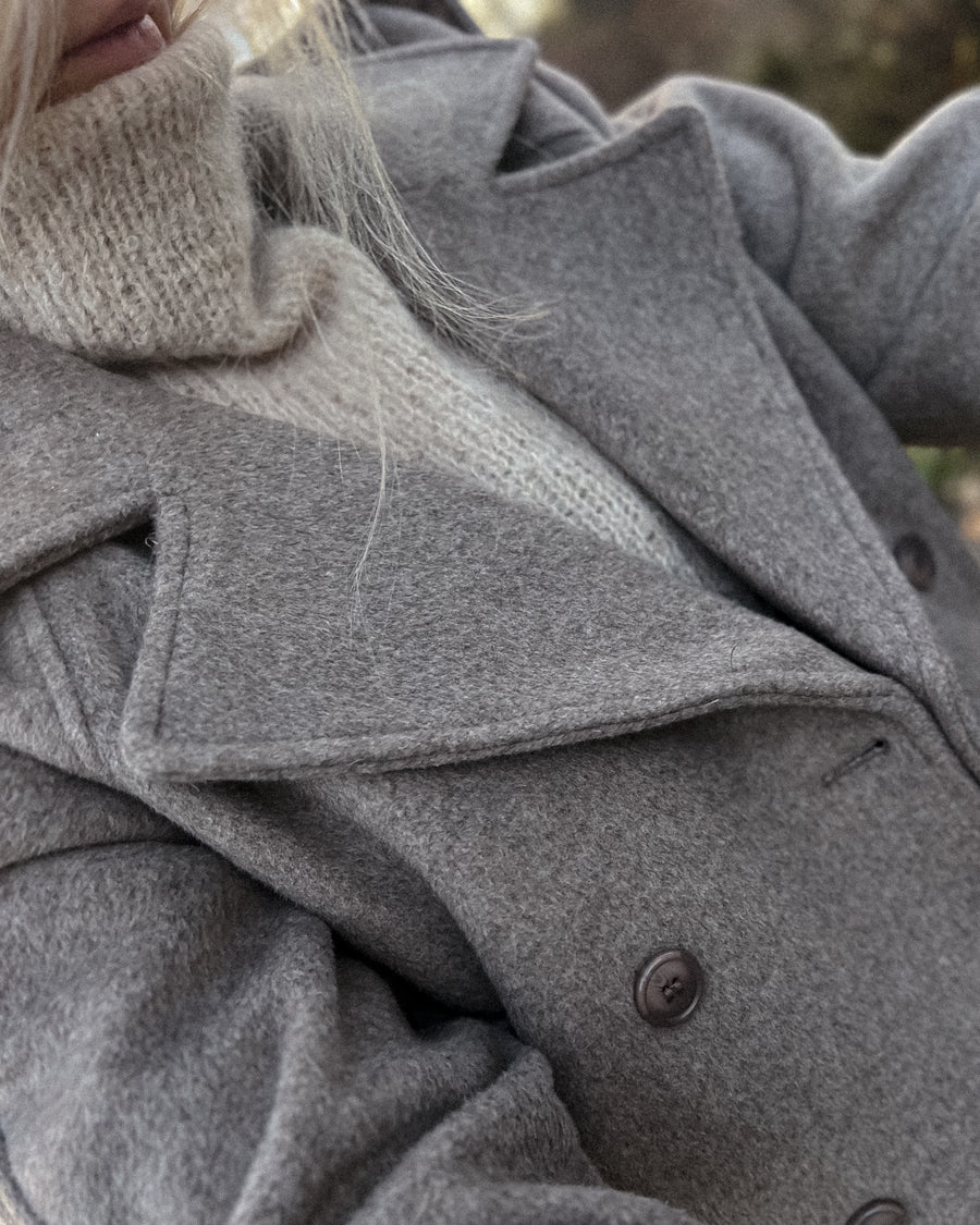 Høj hals på Latte Luxe Sweater i creme, under en grå uldfrakke · strikkes oppefra og ned med udtagninger fra halsen