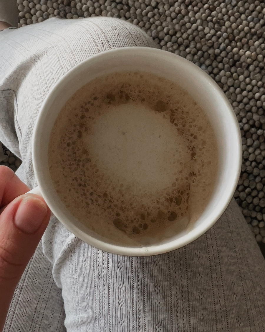 En hånd holder en hvit kopp kaffe over et fang i lys strikk.
