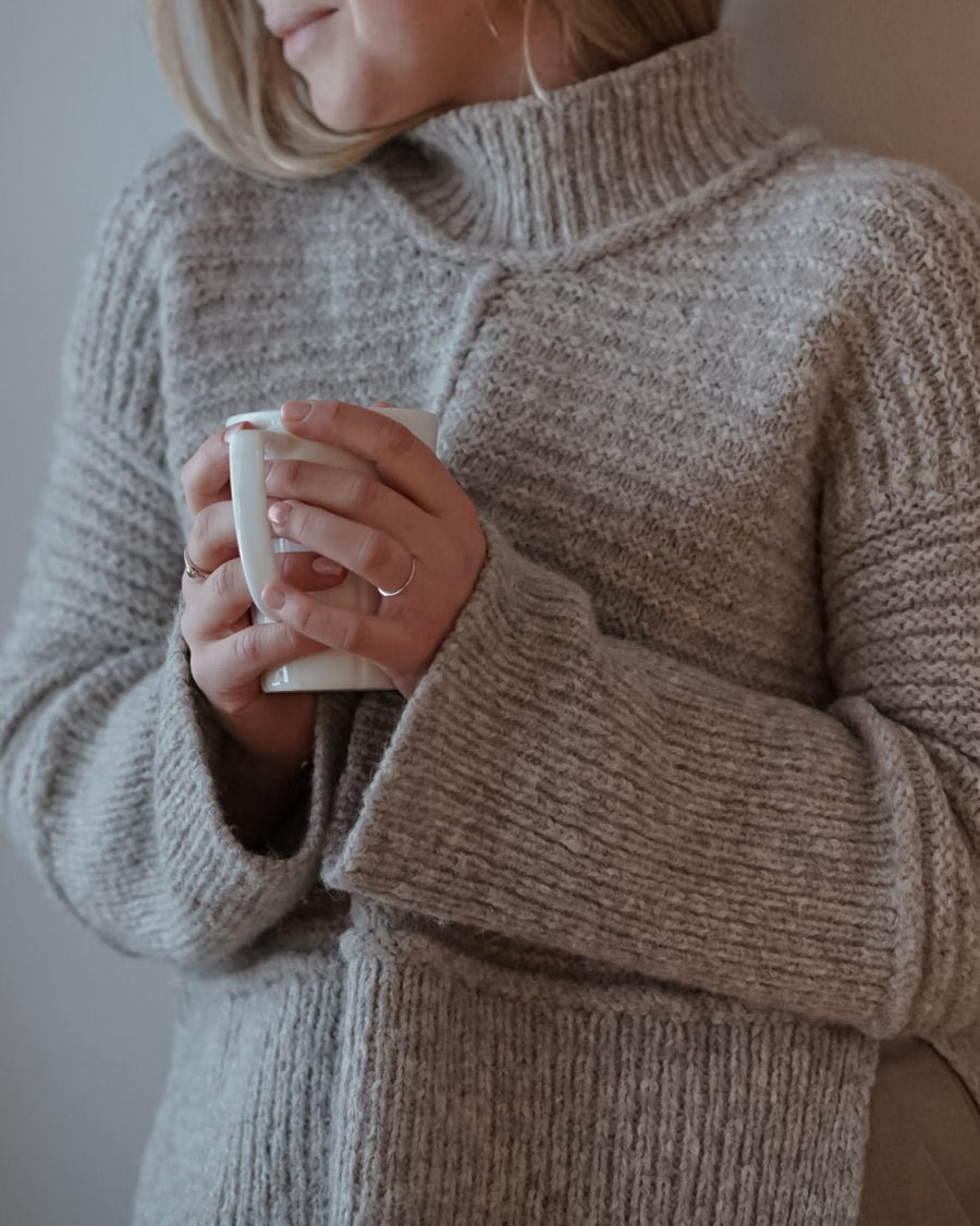 Grå Dawn Sweater med høj hals og ribbet ærme, mens hænderne holder en hvid kop · nedfaldende skulder