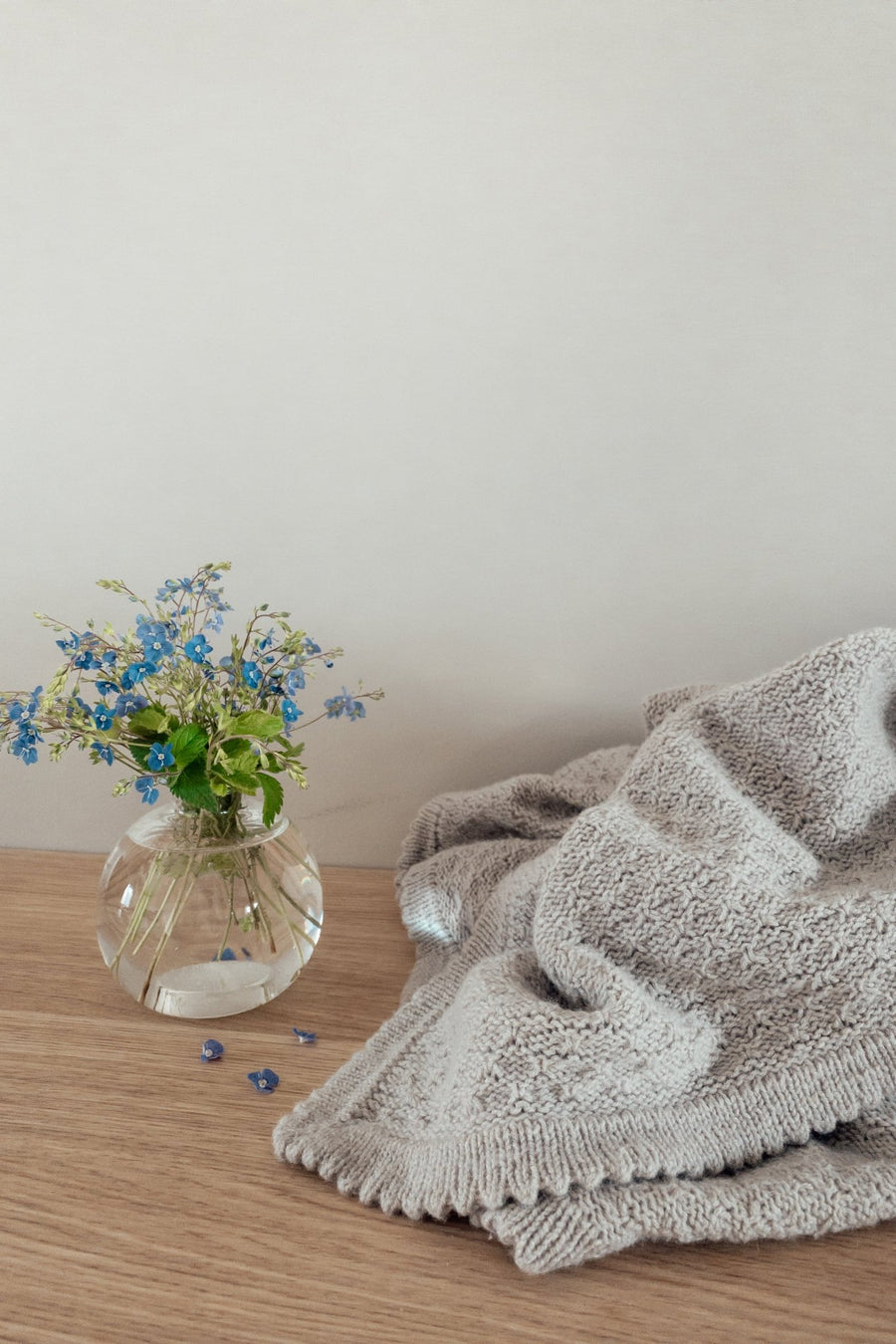 Gråt Granola-tæppe på et træbord ved en vase med blå blomster · picotkanten hækles til sidst hele vejen rundt